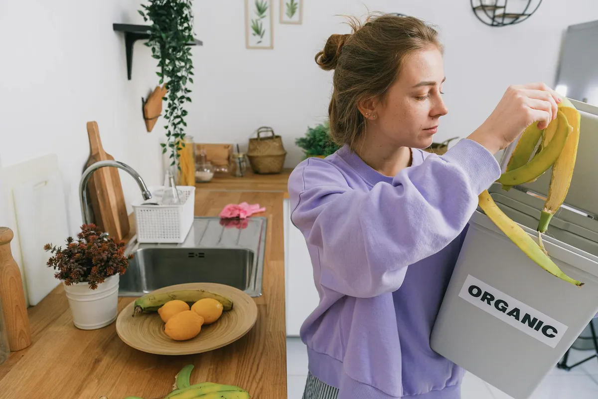 Hand dropping a banana peel into a small countertop kitchen compost bin indoors